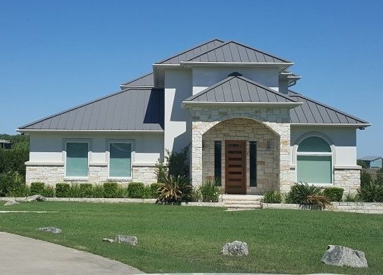 Single-story house with gray metal roof, white exterior, arched stone entry, and green lawn.