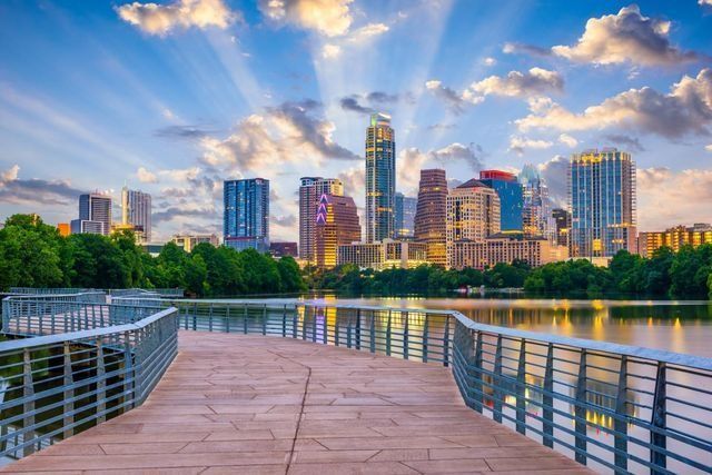 Curved boardwalk beside a calm river with a city skyline at sunset and colorful clouds