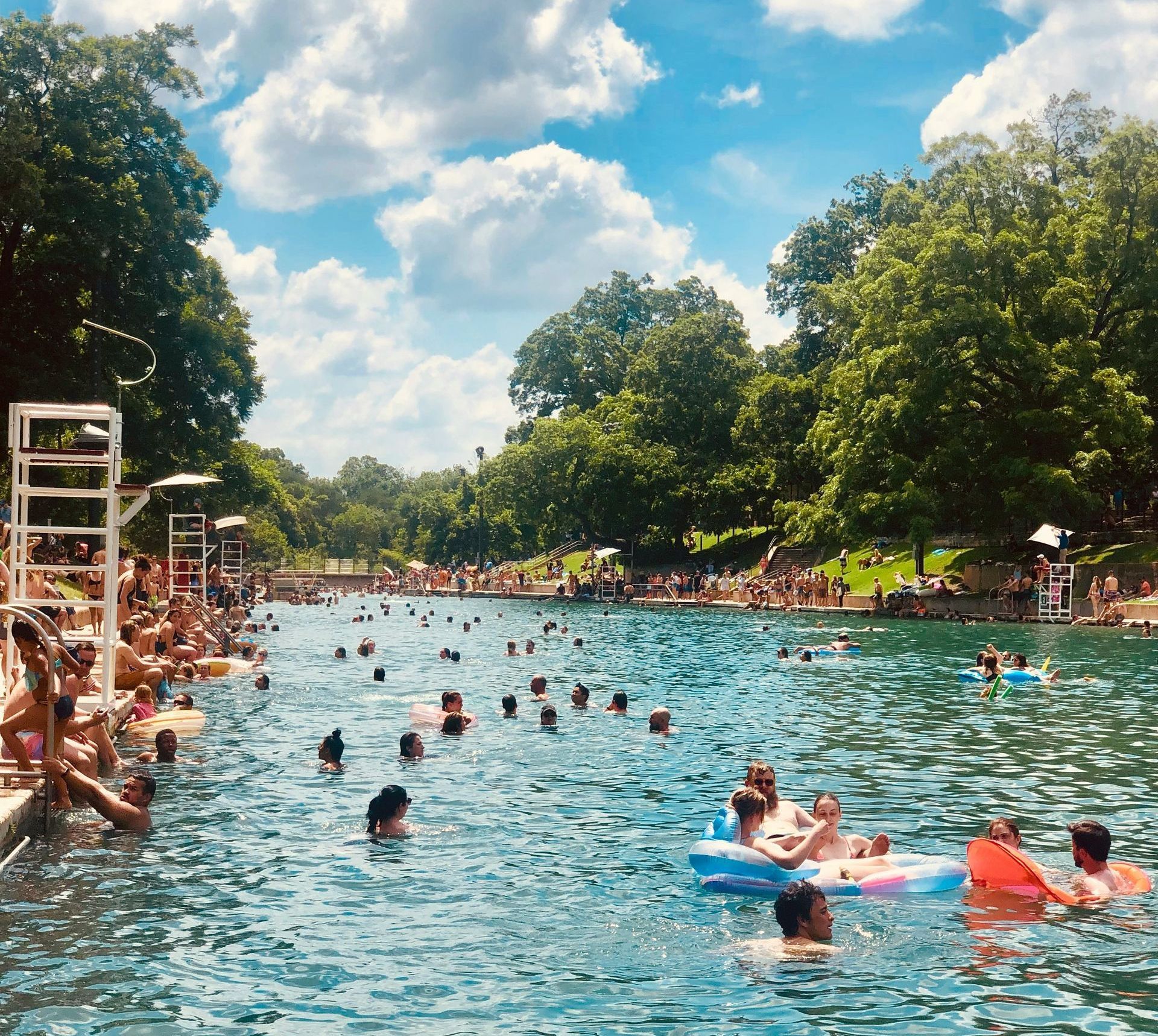 Crowded outdoor pool with swimmers, floaties, and trees under a blue, partly cloudy sky