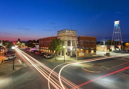 Downtown street at dusk with light trails, historic brick building, and a water tower under a blue sky