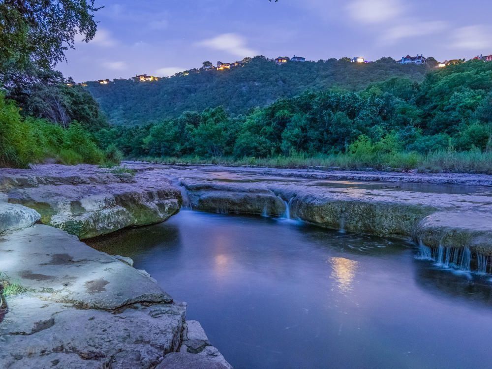 Rocky river pool at dusk with dense green forest and faint lights on the ridge