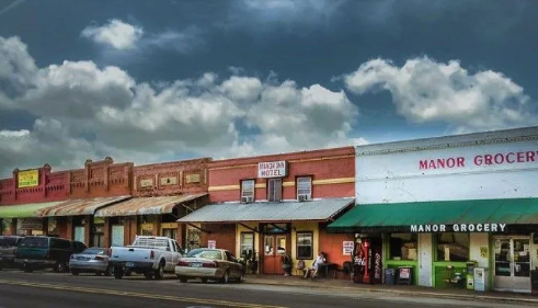 Row of storefronts with parked cars, including Manor Grocery under a white awning