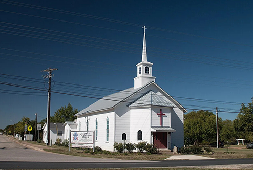 Small white church with a steeple and red door under a clear blue sky beside a road