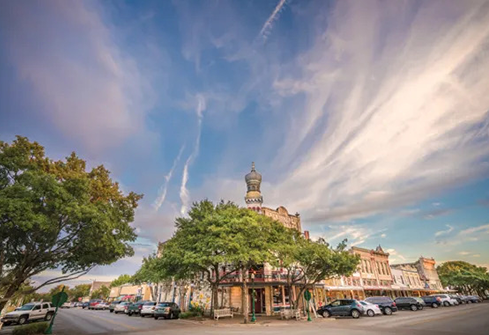 Historic street corner with a tree-lined plaza, parked cars, and dramatic cloudy sky at sunset