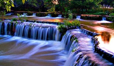 Small cascading waterfalls over rocks in a lush forest, with smooth flowing water and warm sunset light.