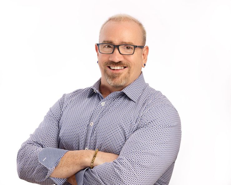 Smiling man with glasses and folded arms, wearing a patterned button-up shirt on a white background