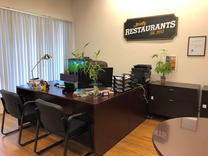 Small office with a wooden desk, black chairs, plants, file cabinets, and a “Restaurants” wall sign.