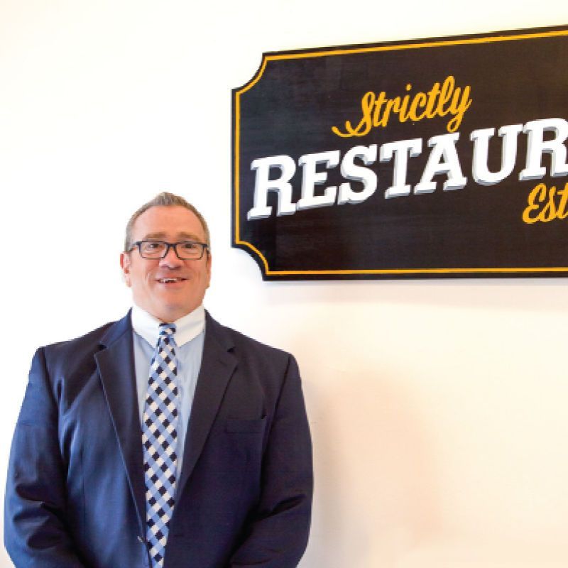 Man in a navy suit standing beside a black and gold restaurant sign on a white wall
