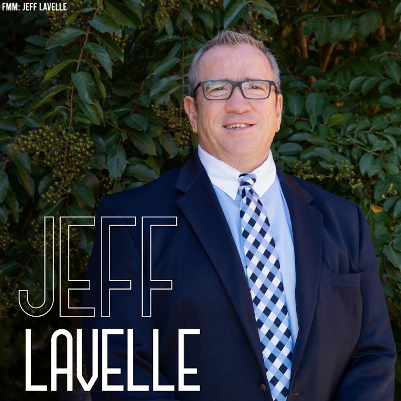 Jeff Lavelle smiling in a dark suit and blue checkered tie against leafy greenery.