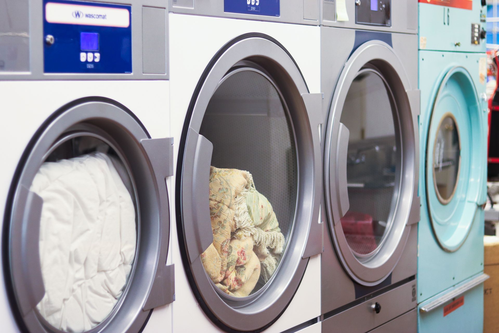 Laundry machines in a laundromat; two with open doors, filled with white and patterned clothes.