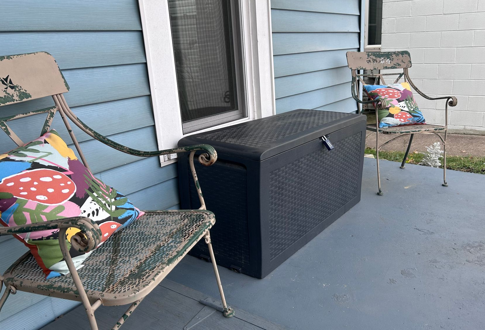 Two metal chairs with colorful pillows flank a black storage chest on a blue porch next to a house.