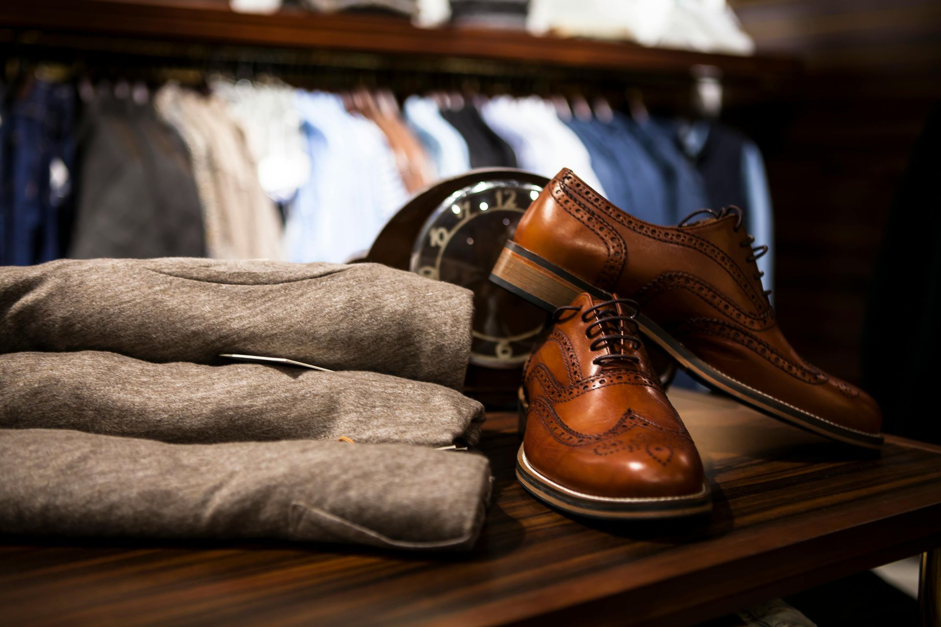 Brown leather dress shoes beside folded gray sweaters on a dark wooden table in a clothing store.