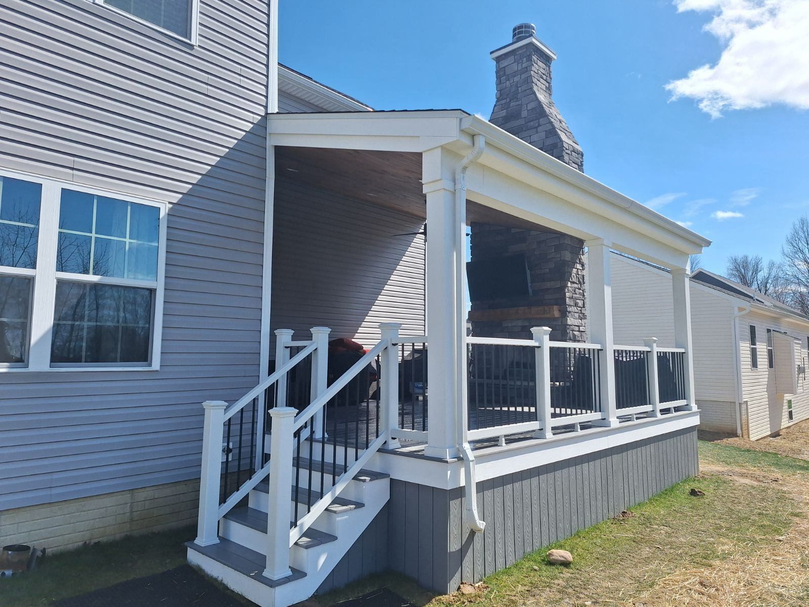 Covered deck attached to a gray house, with steps, white railings, and a brick chimney.