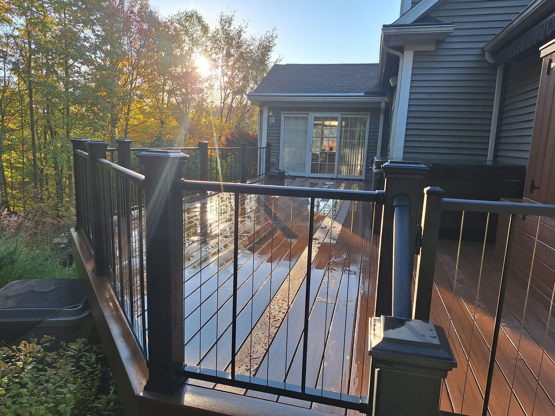 Composite deck with black cable railing and brown boards, sun shining, next to a house.