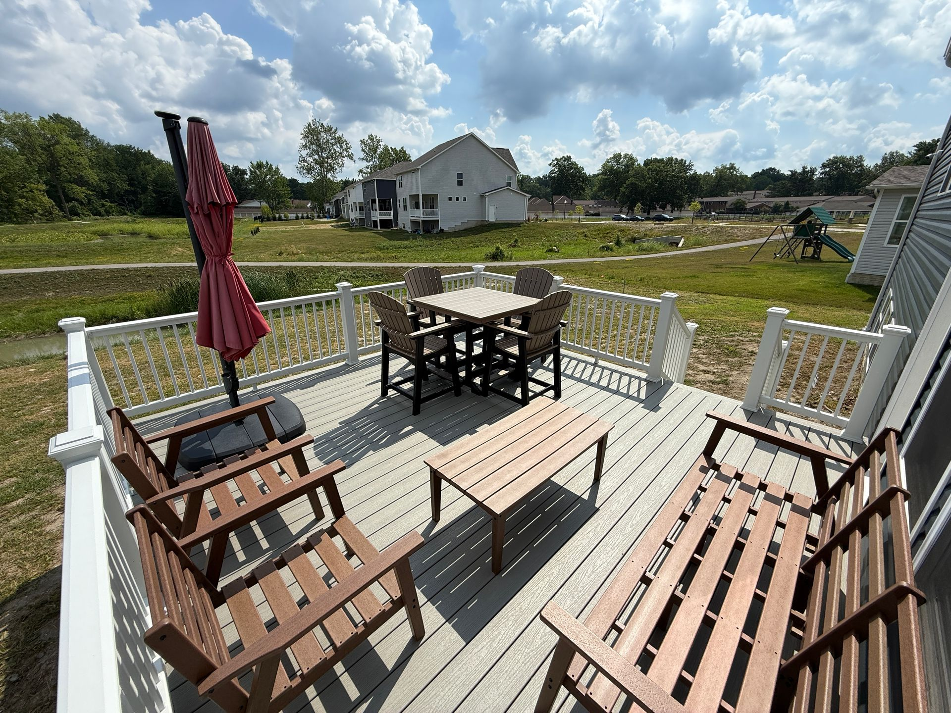 Composite Trex deck with patio furniture, red umbrella, and view of a grassy area, houses, and cloudy sky.