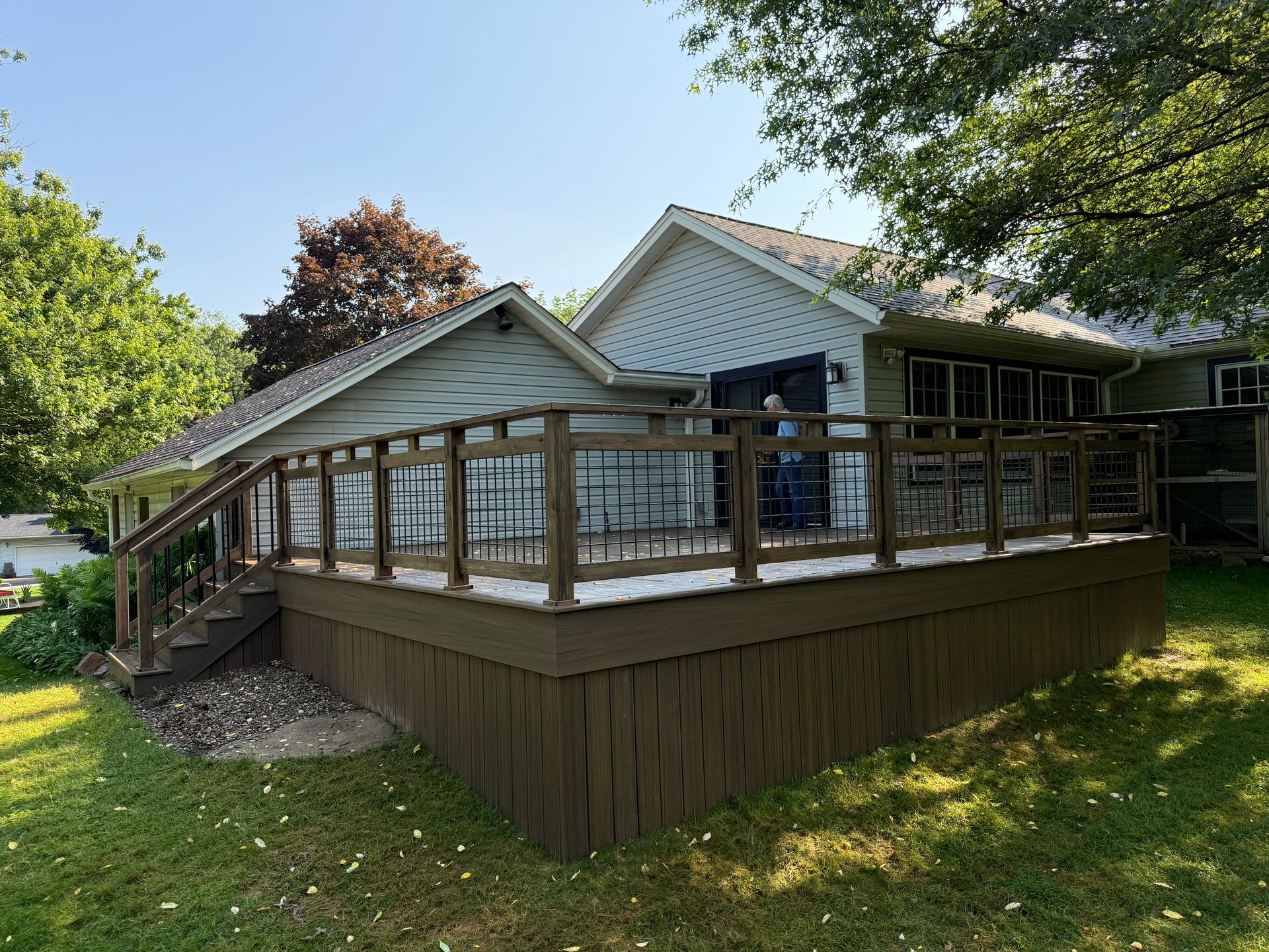 Brown deck with hog wire panels attached to a light green house, surrounded by green grass and trees.