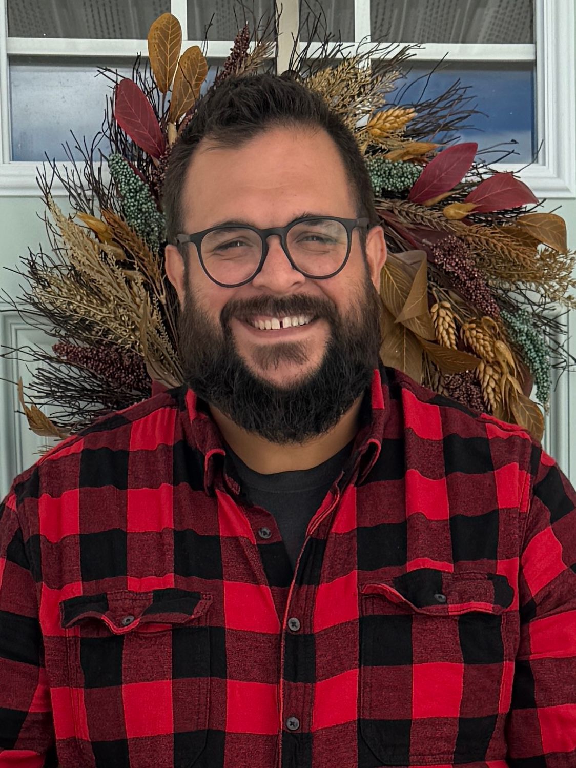 Man with a full beard wearing glasses and a red and black plaid shirt smiles in front of a fall wreath.