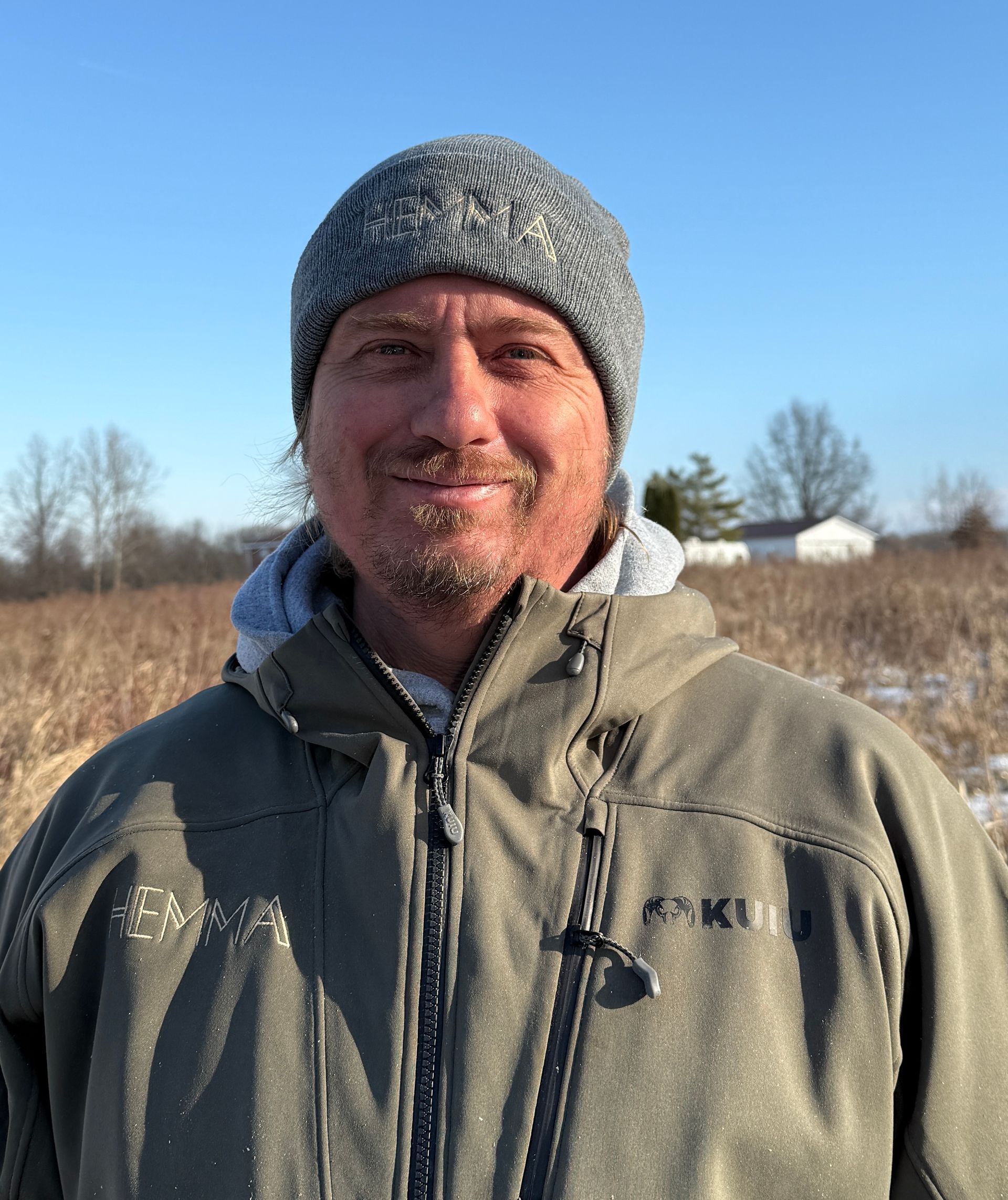Man in gray beanie and jacket smiles outdoors in field.
