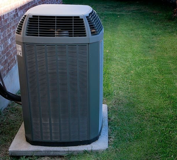 Gray air conditioning unit on a concrete pad, set on grass next to a brick wall.