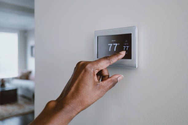 A hand adjusting a digital thermostat displaying 77 degrees, mounted on a white wall indoors.