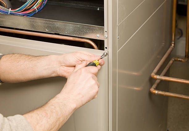 Person using a screwdriver to remove a panel on a furnace. Copper pipes and wires visible.