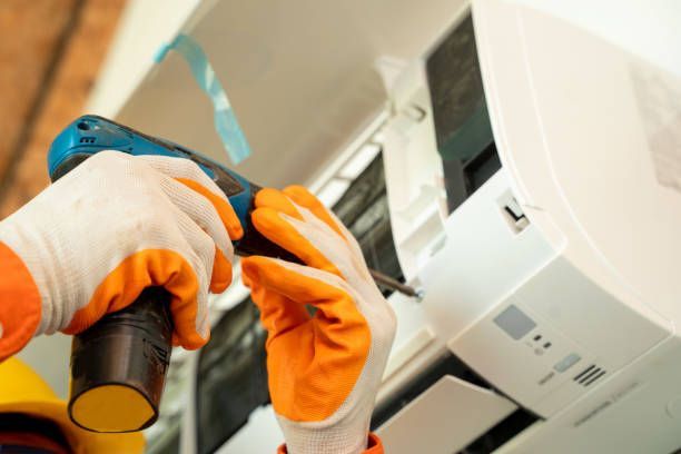 Person in orange gloves uses a power drill to install an AC unit on a wall.