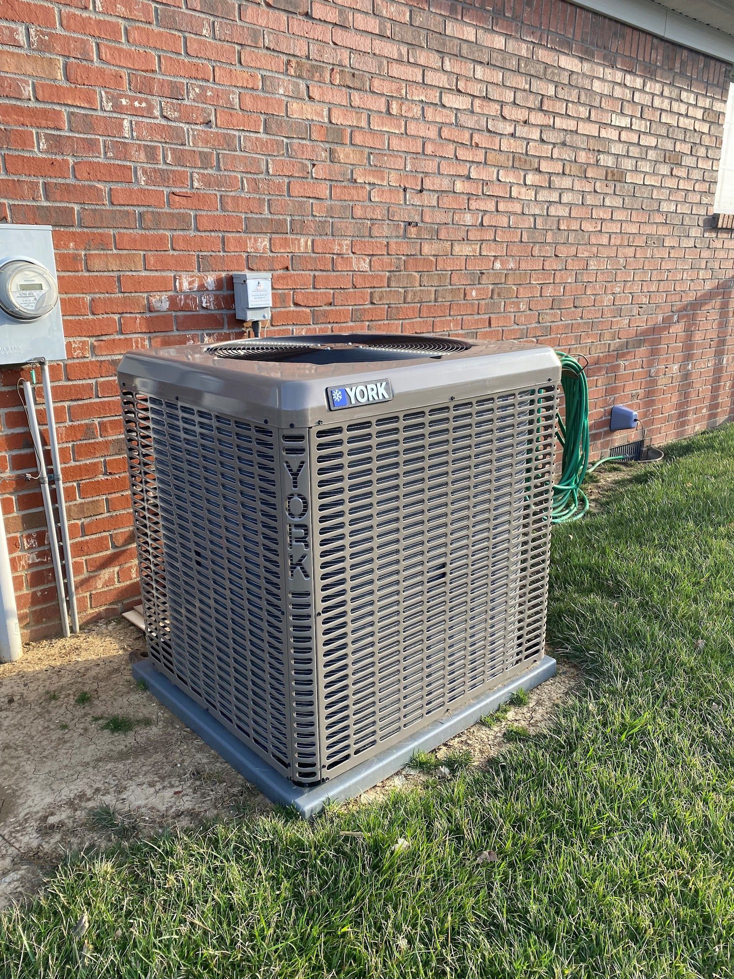 A tan York air conditioning unit sits on a concrete pad against a red brick exterior wall near a green lawn.