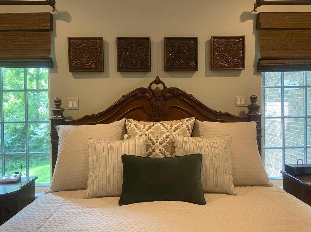 Elegant bedroom with carved wooden headboard, white bedding, and green accent pillow between two windows.