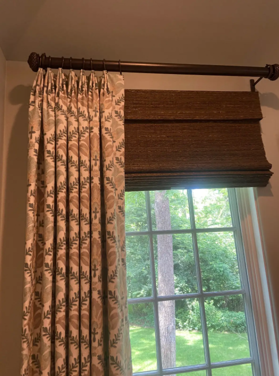 Floral curtain and brown Roman shade on a window with green trees outside
