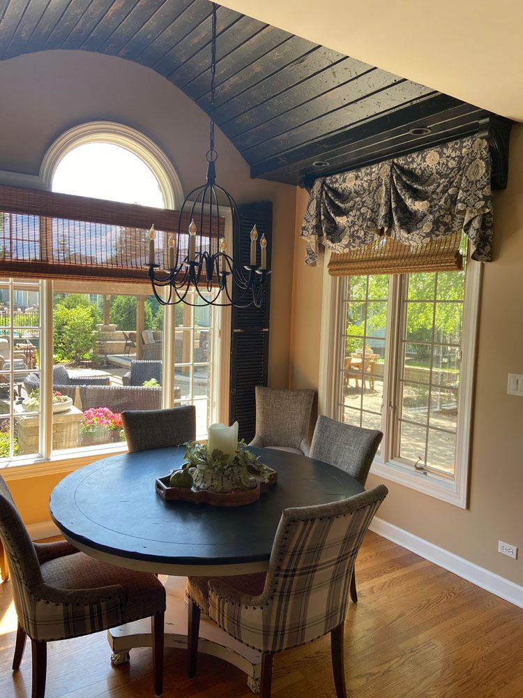 Dining nook with round black table, woven chairs, large windows, and vaulted wood ceiling in warm sunlight