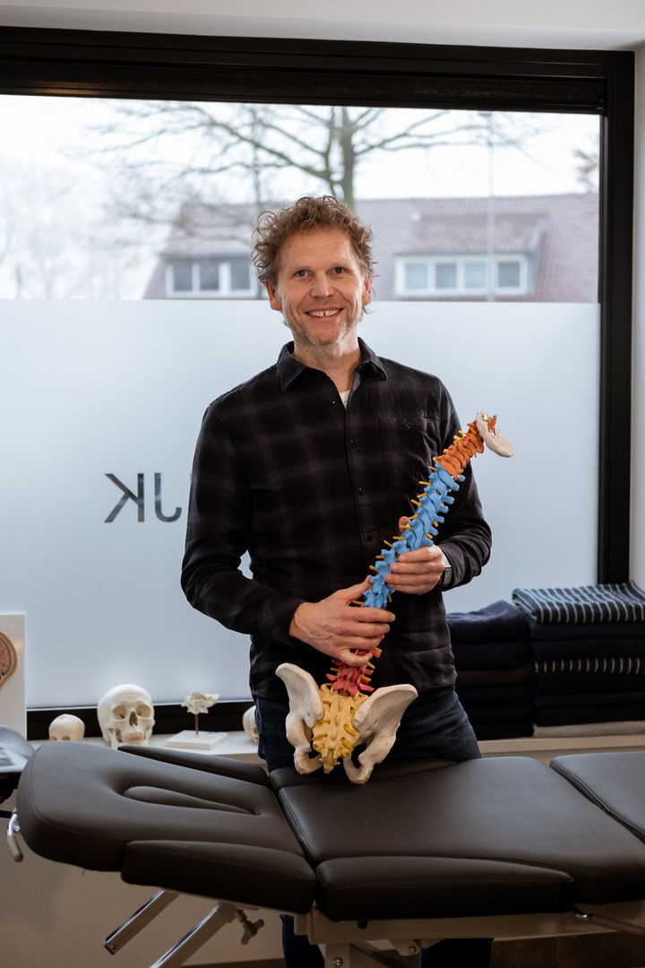 Een man houdt een kleurrijk skeletmodel vast naast een massagetafel in een lichte behandelkamer.