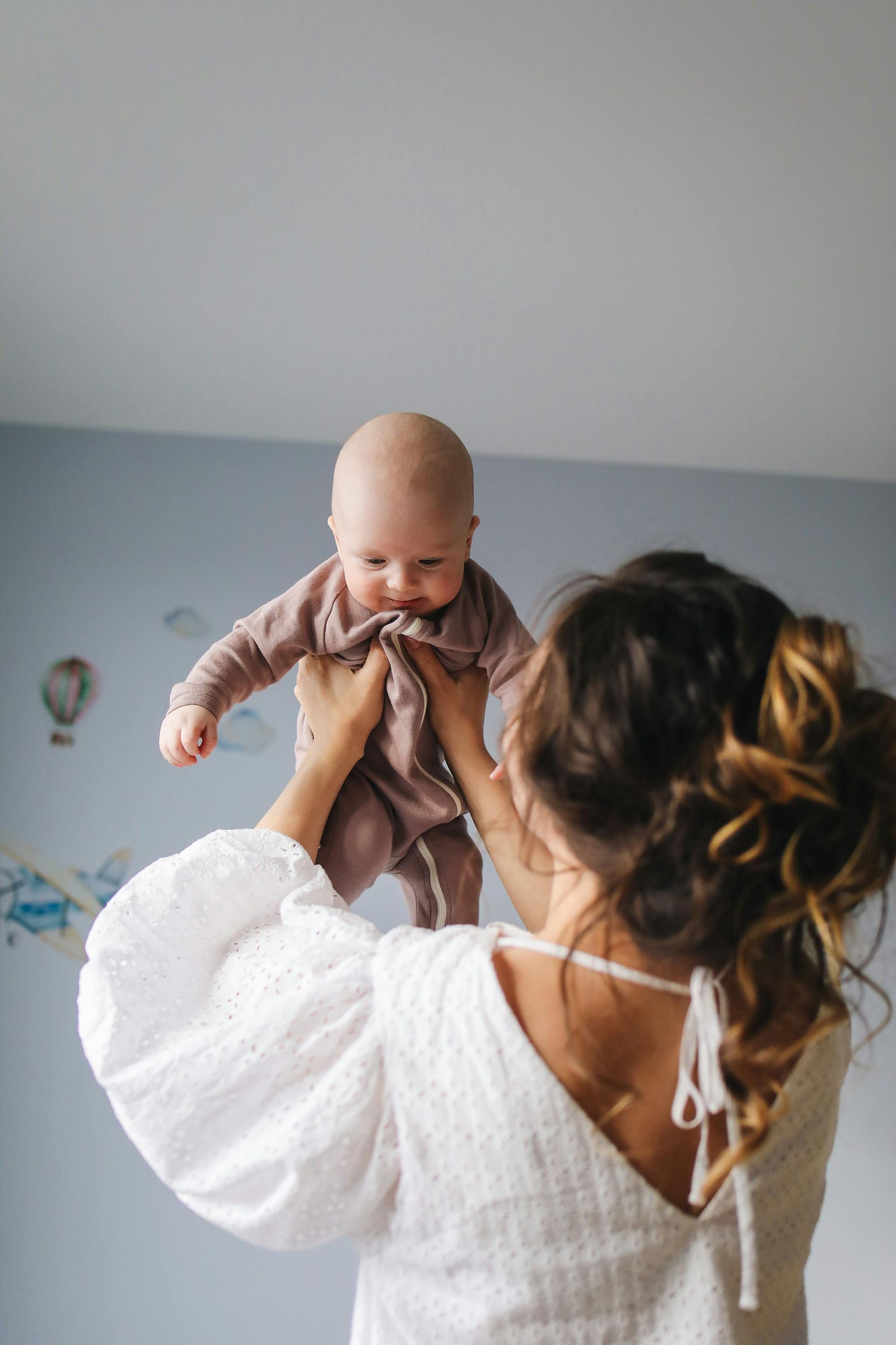 Een vrouw tilt een baby op in een kamer, beiden gekleed in witte kleding, tegen een lichtgekleurde muur.