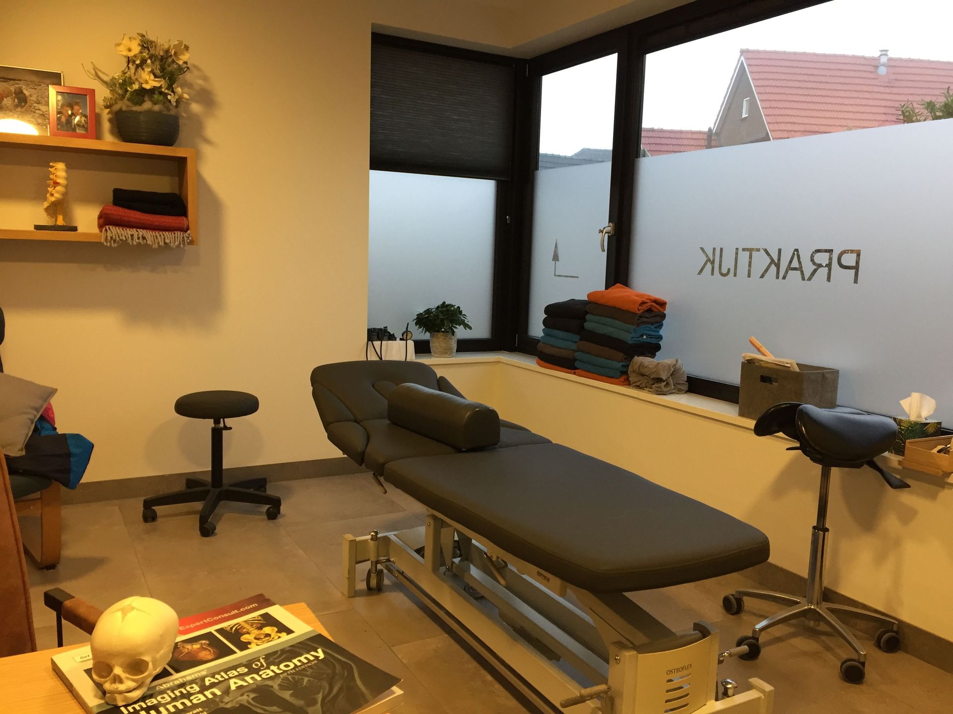 Massageruimte met twee behandeltafels, krukken en grote ramen; op het bord staat 