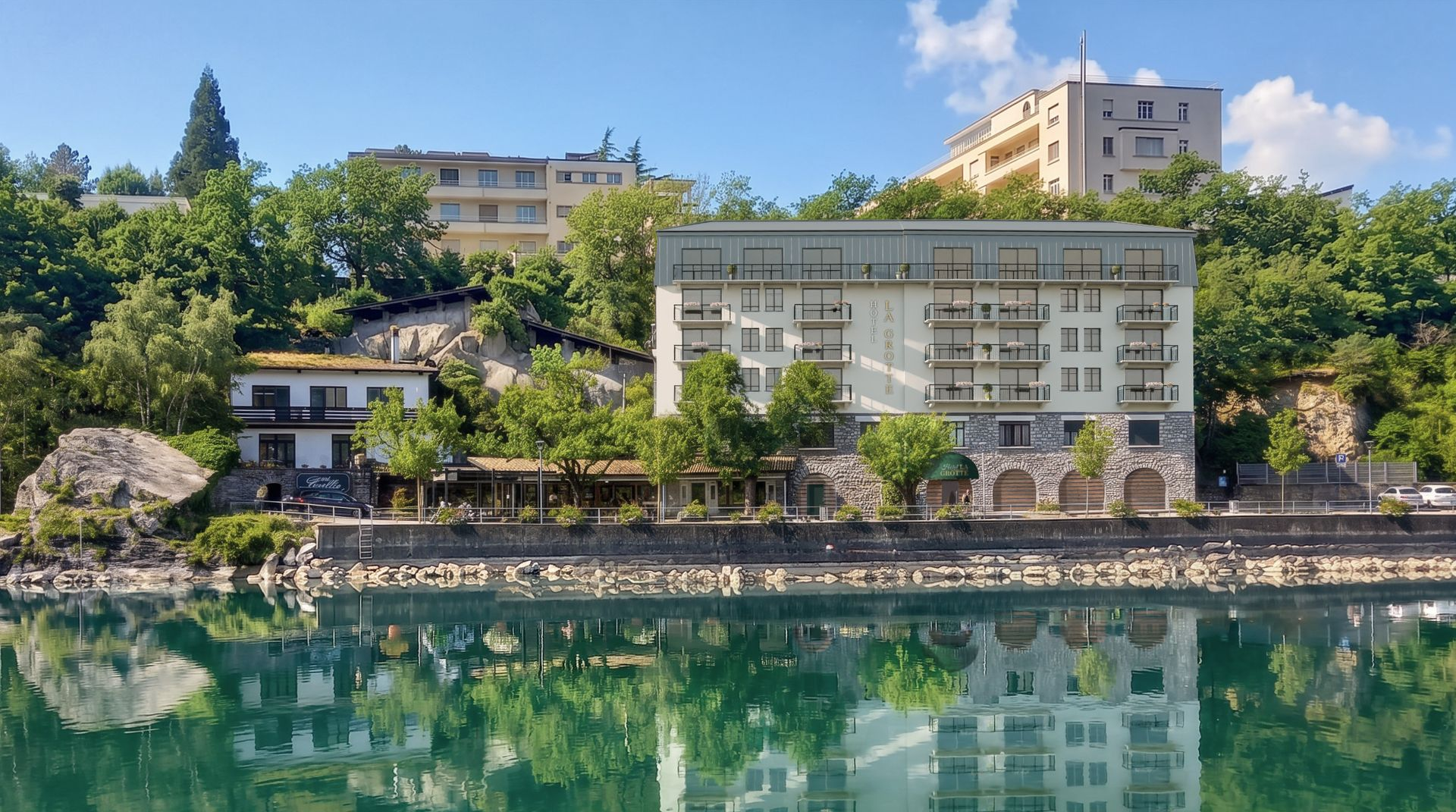 Immeuble d'appartements en bord de mer se reflétant dans une eau turquoise calme, entouré d'arbres et de collines.