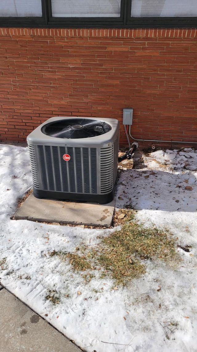 Outdoor air conditioner unit beside a brick wall, with snow patches on the ground.