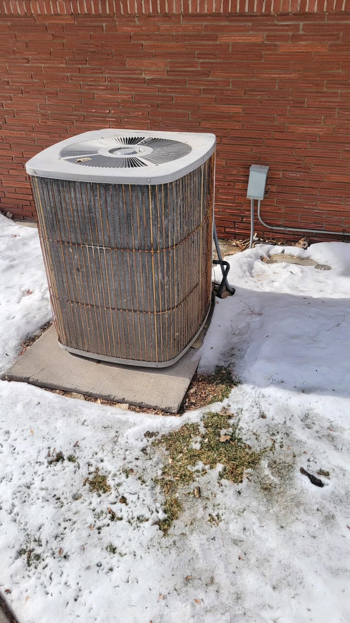 Outdoor air conditioner unit beside a brick wall, surrounded by snow.