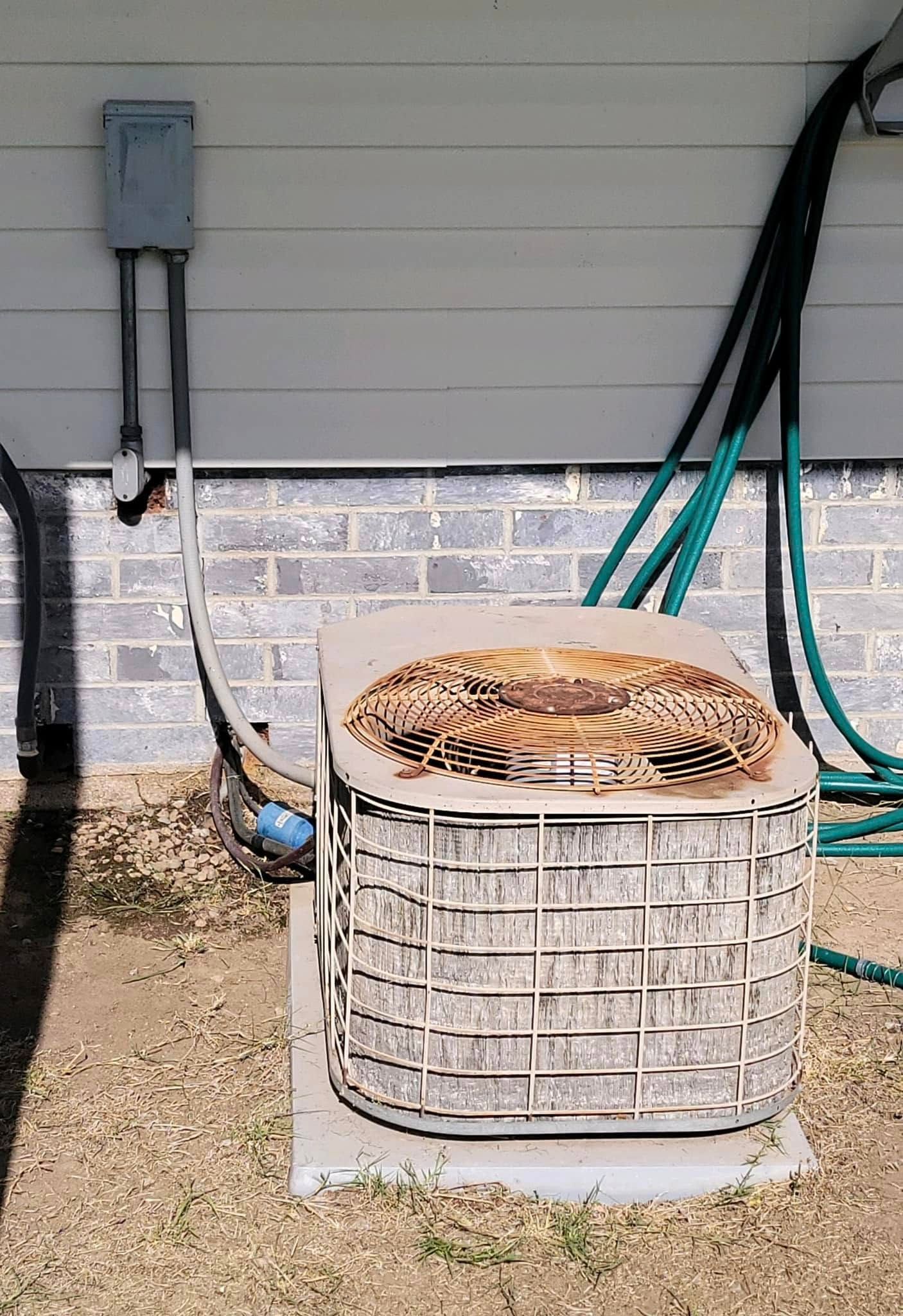 Outdoor HVAC unit beside a house wall with hoses and a wall-mounted electrical box