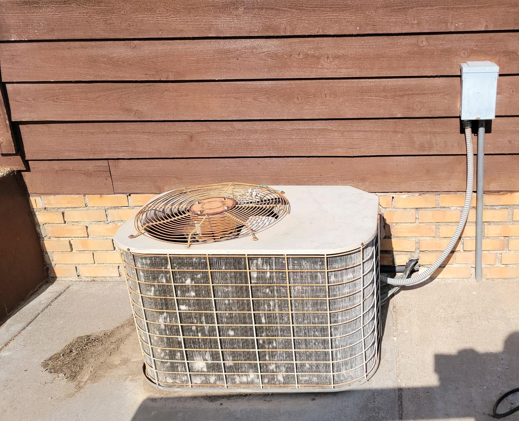 Outdoor HVAC unit against a wooden wall, with a rusty fan cover and a small electrical box nearby