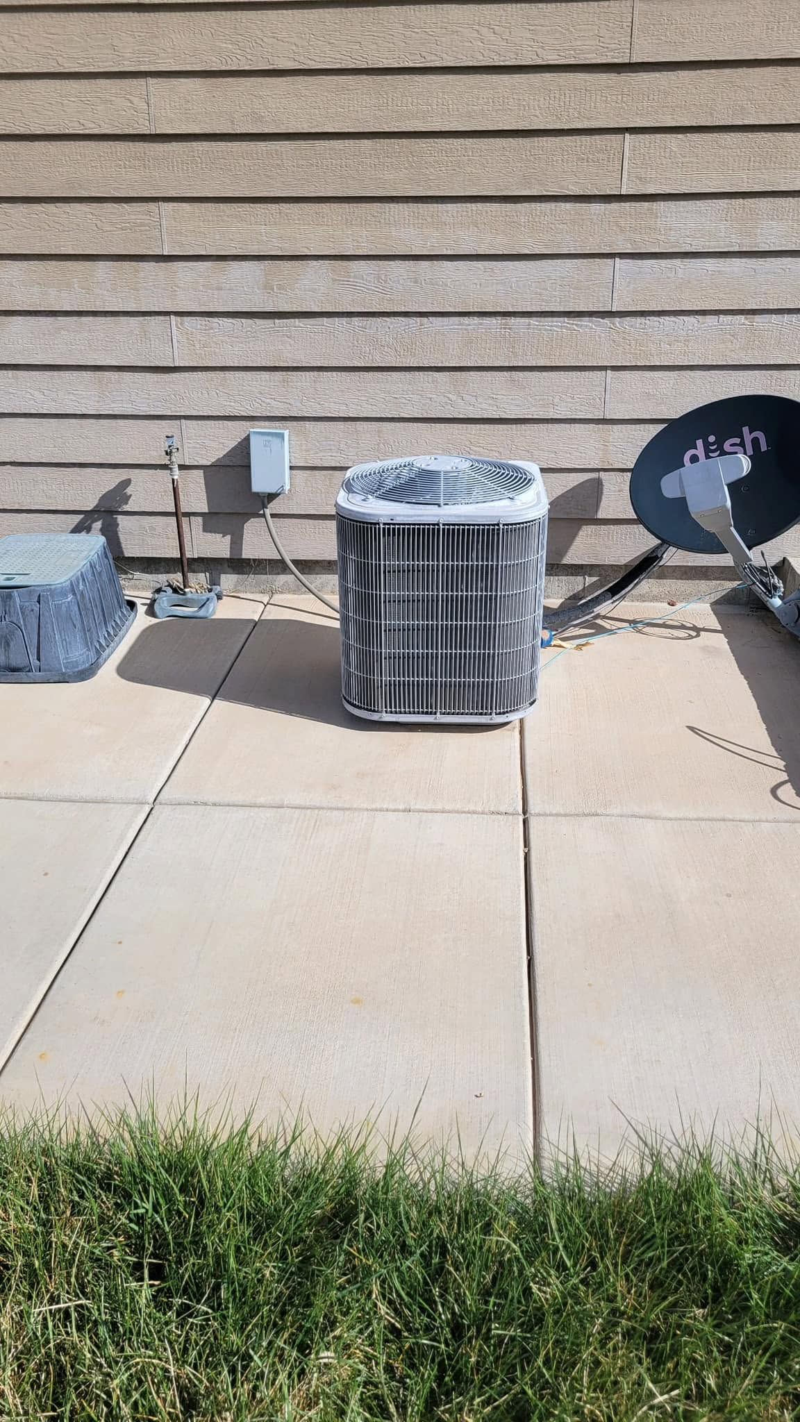 Outdoor HVAC unit beside a brick wall, with a grill on the right and grass in the foreground.