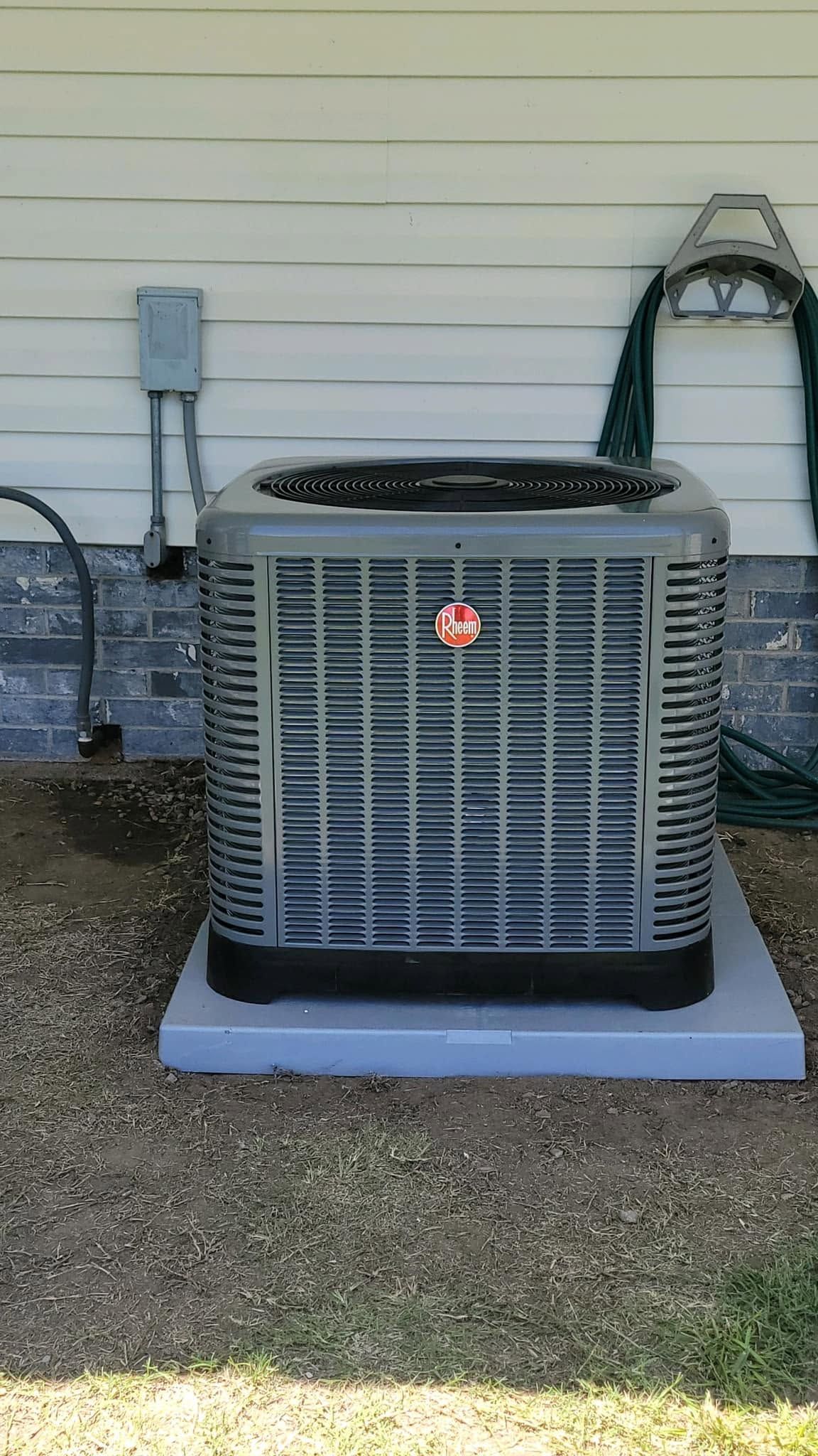 Gray outdoor air conditioner unit beside a house wall on a concrete pad with hoses attached