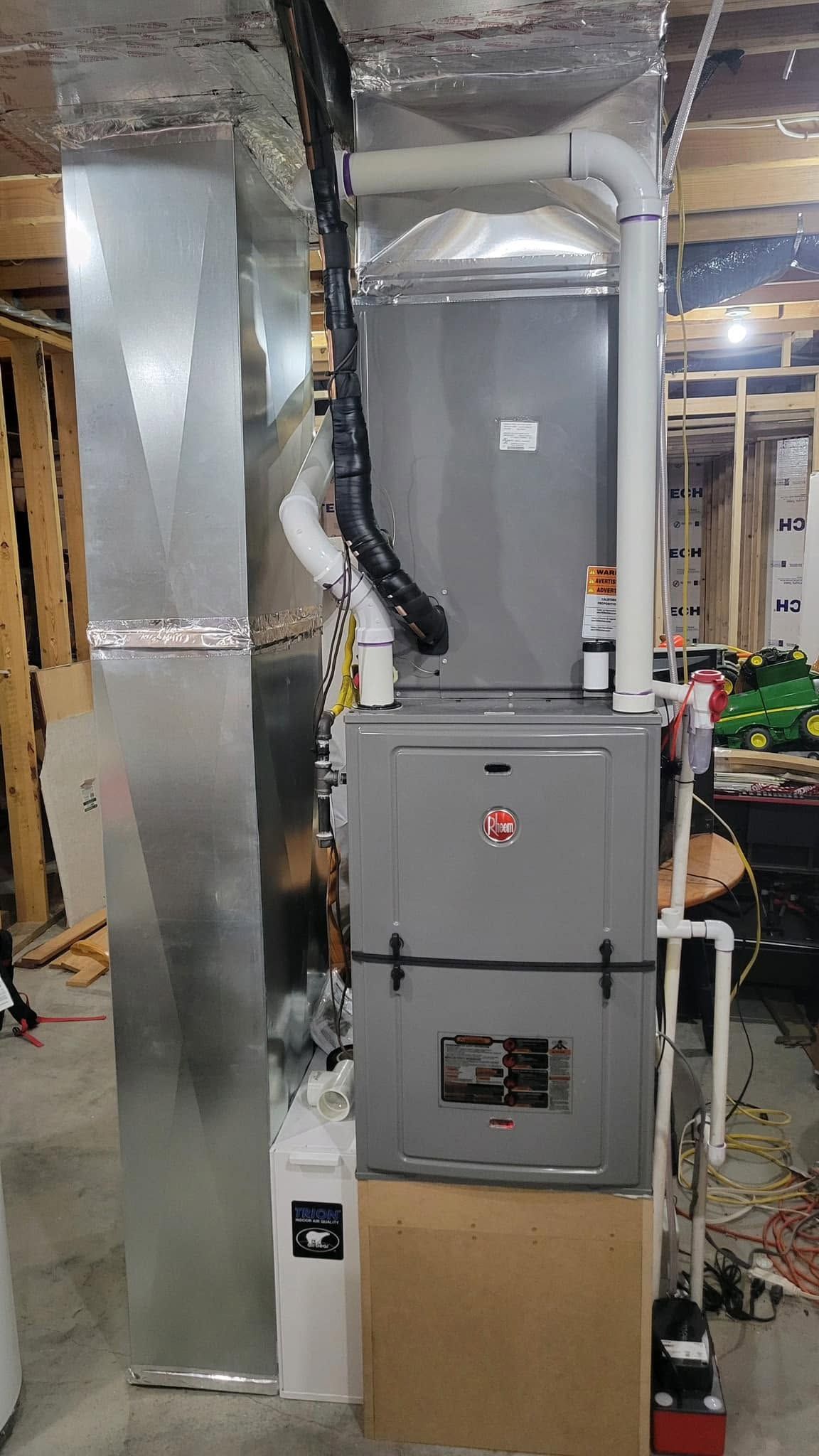 Gray furnace unit in a basement with silver ductwork and exposed beams, surrounded by utility equipment.