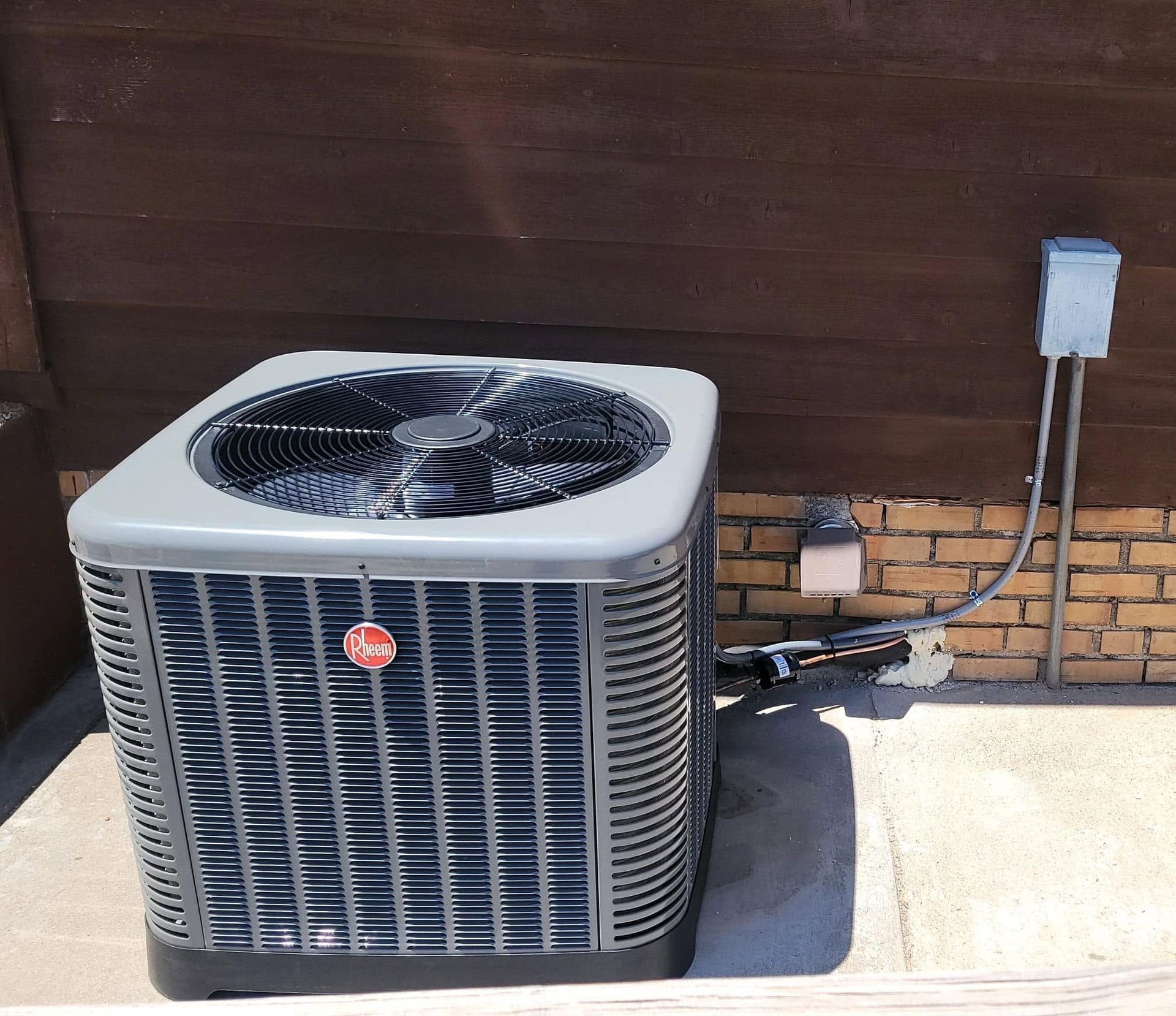 Outdoor air conditioner unit beside a wooden fence and brick wall, with electrical conduit on the right.