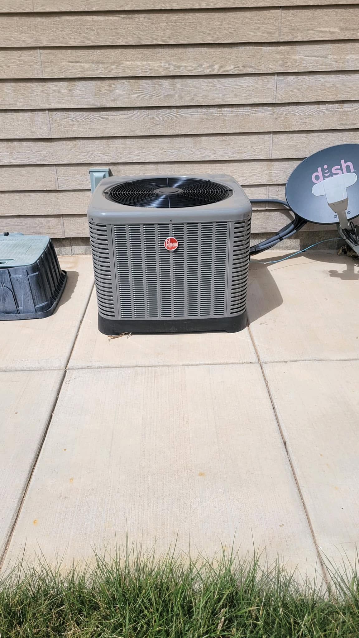 Outdoor air conditioner unit on a concrete patio beside a beige house wall and grass border