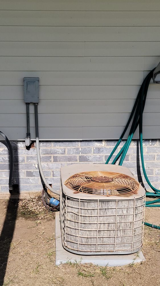 Outdoor air conditioner unit beside house wall with electrical box, cables, and green hose attached