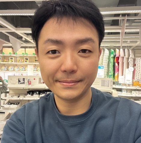 一名男子在室內微笑,可能是在商店裡。他有著深色頭髮,身穿藍色運動衫。他身後是貨架。