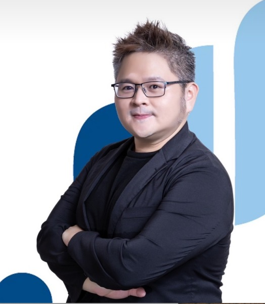 戴眼鏡的男子,雙手交叉,面帶微笑。身穿黑色西裝外套和襯衫。背景為白色和藍色。