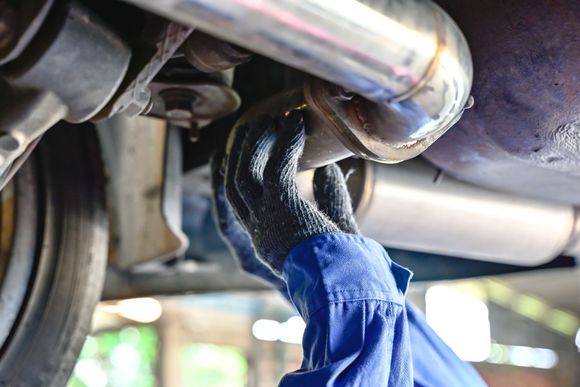 Mechanic in blue coveralls, wearing gloves, working on a car exhaust system underneath a vehicle.