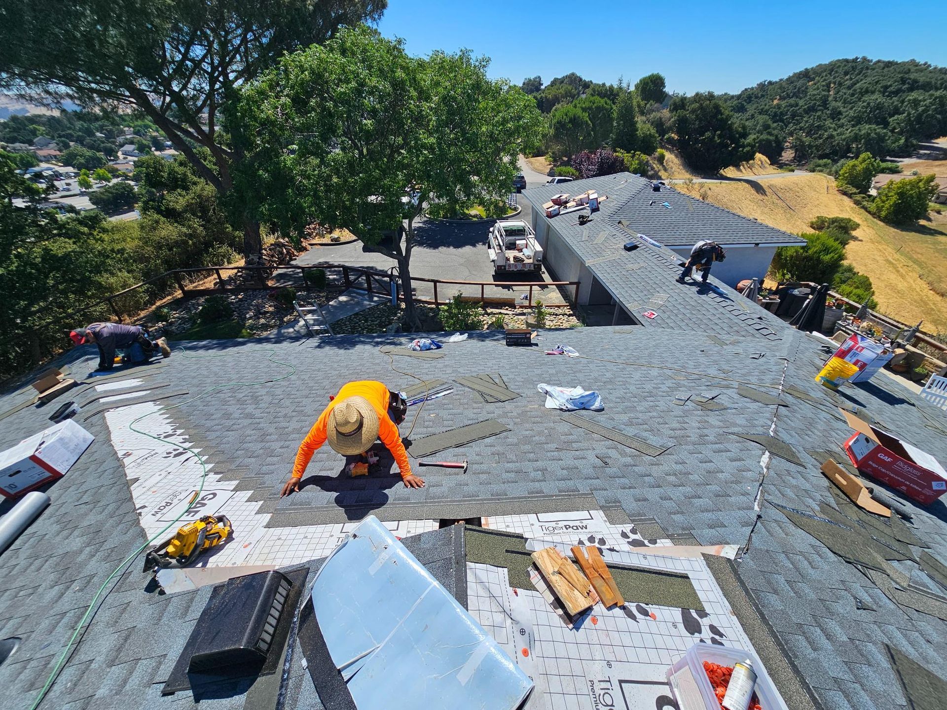 Workers repairing a gray shingle roof on a sunny hillside home, with trees and a road below