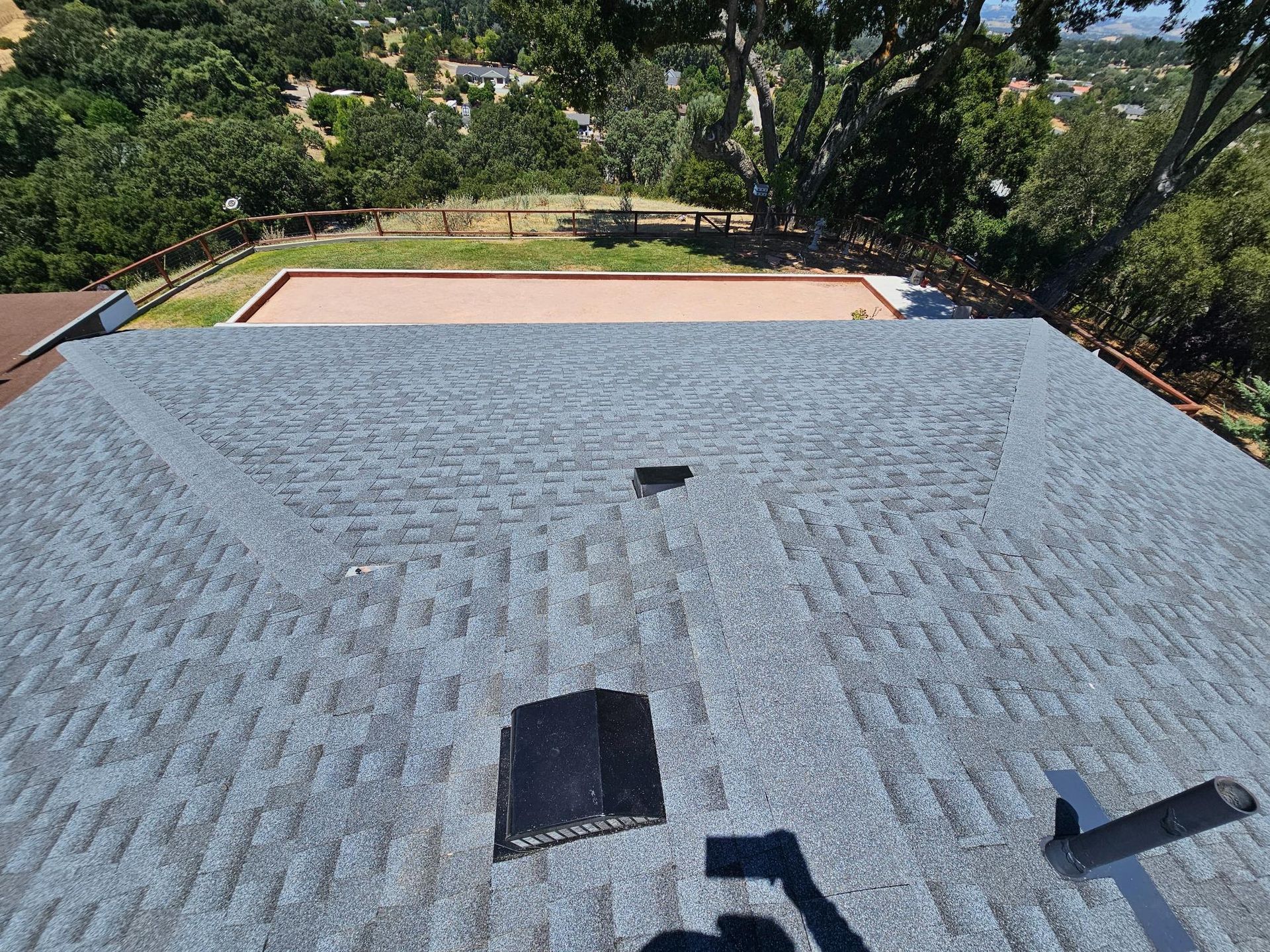 Roof with gray shingles in foreground, overlooking a backyard with trees and a small patio or pool area.