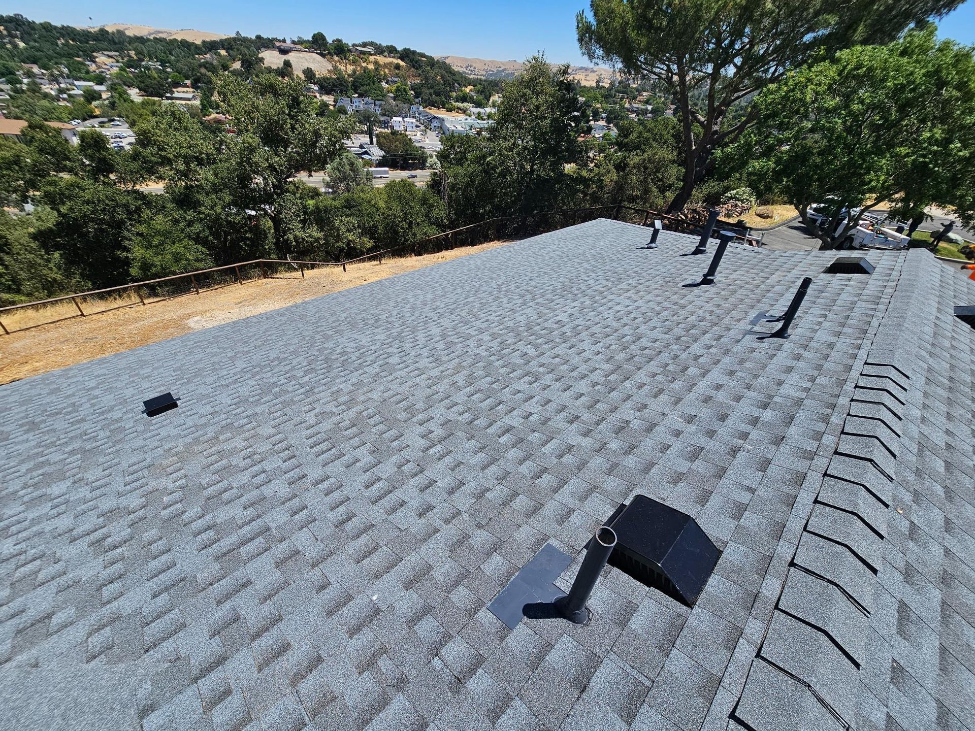 Gray shingle roof with vents and trees in the background on a sunny hillside